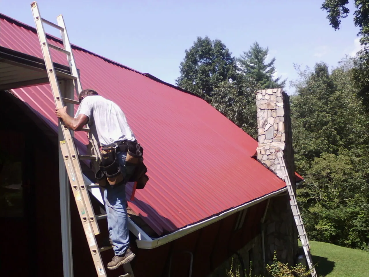 Roofer on a ladder beside a red metal roof and stone chimney — Soffit & Fascia Repair by Roofing Brothers in Semmes